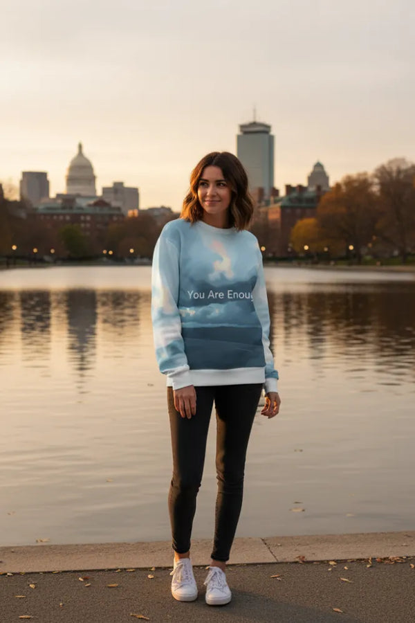 Woman wearing a sweater with text standing by a lake with a city skyline in the background