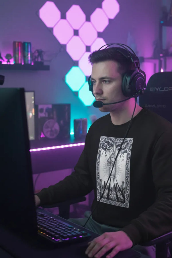 Person wearing headphones and a black sweatshirt with a graphic design, sitting in front of a computer in a room with colorful lighting.