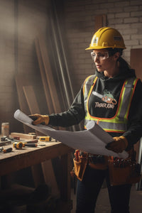 Person in a construction setting wearing a hard hat and safety glasses, holding blueprints.