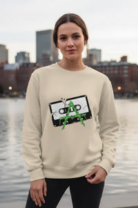 Person wearing a beige sweatshirt with a graphic design, standing by a waterfront with buildings in the background.