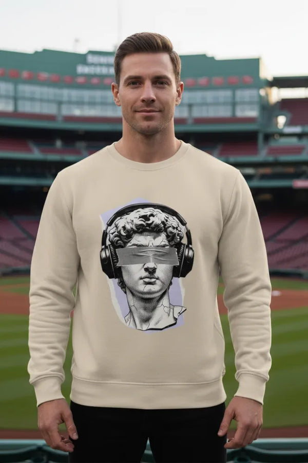 Man wearing a beige sweatshirt with a graphic design of a classical statue wearing headphones in front of a stadium.