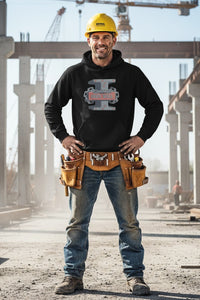 Man in a construction site wearing a black hoodie with a logo, blue jeans, and a yellow hard hat.