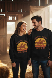 Two people wearing matching black sweatshirts with a sunset design and text in a barn setting.