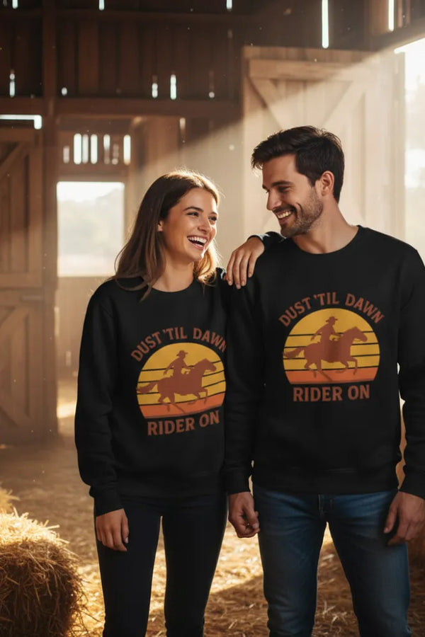 Two people wearing matching black sweatshirts with a sunset design and text in a barn setting.